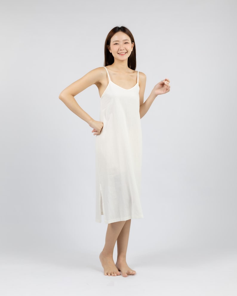 May include: A woman wearing a white sleeveless midi dress with a relaxed fit. She is smiling and standing on a white background.