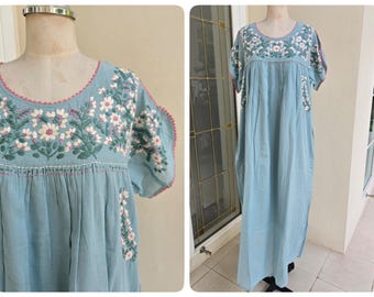 May include: A light blue, floor-length dress with short sleeves. The dress features intricate white and pink floral embroidery around the neckline and sleeves. The dress is displayed on a mannequin.
