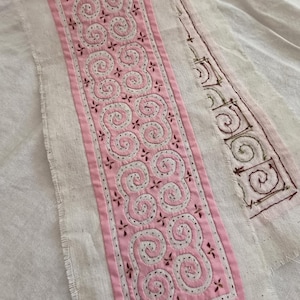 Puede incluir: Una tira de tela color hueso con un panel central rosa decorado con patrones de espirales bordados en blanco y pequeños detalles de punto de cruz en rojo oscuro. A la derecha, una sección separada de tela muestra una cuadrícula dibujada con tinta roja.