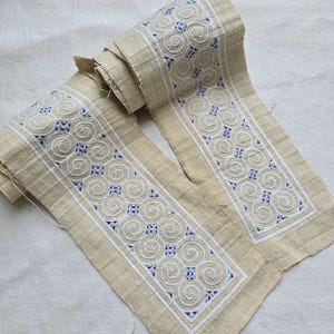 Puede incluir: Dos tiras de tela beige con bordados de espirales y patrones geométricos blancos, azules y verdes. La tela tiene un aspecto crudo y texturizado, lo que sugiere una calidad artesanal. El diseño está centrado en la tela.