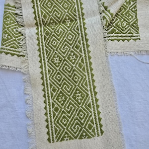 May include: Close-up of a cream-coloured fabric with a geometric green embroidered design. The design features repeating diamond and square patterns. The edges of the fabric are frayed, giving it a rustic look. The fabric is likely used for decorative purposes.