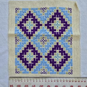 May include: A square piece of fabric with a detailed cross-stitch pattern. The design features repeating geometric shapes in shades of purple, light blue, and green, set against a beige background. A ruler is placed along the bottom edge.