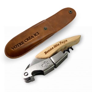Könnte beinhalten: Ein Korkenzieher aus Holz und Metall mit einem braunen Lederetui. Der Korkenzieher hat einen Holzgriff mit dem eingravierten Text "Bonne fête Papa". Das Etui hat den eingravierten Text "VOTRE CREA ICI".