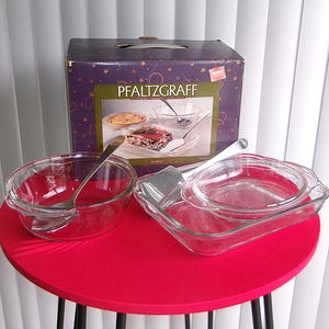 May include: A clear glass Pfaltzgraff bakeware set with a box in the background. The set includes a round bowl with a spoon, a rectangular dish with a lid, and a serving utensil. The box is purple with the Pfaltzgraff logo.
