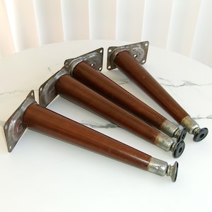 May include: A set of four tapered furniture legs with a dark brown wood finish. Each leg features a rectangular metal mounting plate with four screw holes at the top and a small black circular foot at the bottom for floor protection.