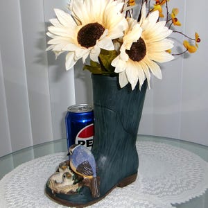 May include: A dark teal ceramic boot vase with a bird and nest design, holding sunflowers and other flowers. A blue Pepsi can is next to the vase. The boot vase is a unique decorative item.