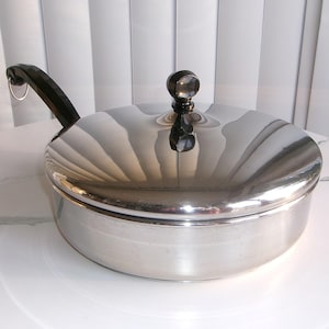 May include: A stainless steel cooking pot with a lid and a black handle. The lid has a black knob. The pot is round and reflective, with the handle extending to the left. The pot is sitting on a white surface.