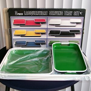 May include: A 3-piece lacquerware serving tray set in a black box with the text 3 PIECE LACQUERWARE SERVING TRAY SET. The set includes rectangular green trays with white edges and integrated handles, partially wrapped in clear plastic packaging.