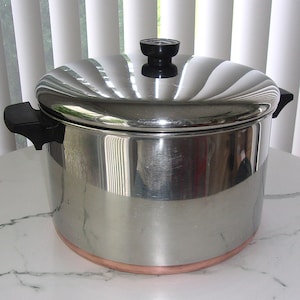 May include: A stainless steel cooking pot with a lid. The pot has a copper-colored band at the base and black handles. The lid has a black knob and a ridged design.