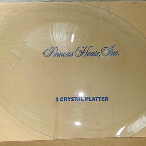 May include: A clear glass platter featuring etched floral accents along the rim. The center of the platter displays the blue text Princess House, Inc. and 1 CRYSTAL PLATTER. The platter is circular with a smooth, reflective surface and transparent finish.