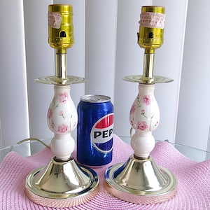 May include: Two vintage table lamps with a white and pink floral design. The lamps have gold-colored bases and tops, and the shades are not included. A blue Pepsi can is next to the lamps.