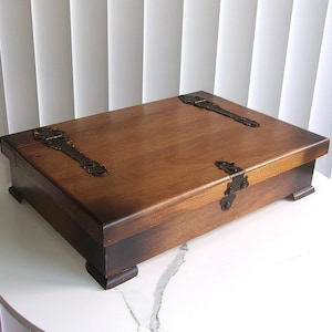 May include: A rectangular wooden box with a dark brown finish, featuring decorative black metal hinges on the top lid and front edge. The box is supported by four small, sturdy feet at the corners, providing a raised profile for this storage container.