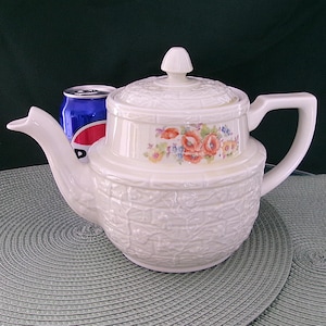May include: A white ceramic teapot with a floral design and a textured basketweave pattern. The teapot has a lid with a knob and a curved handle. The floral design features red, orange, and yellow flowers. A can of soda is next to the teapot.