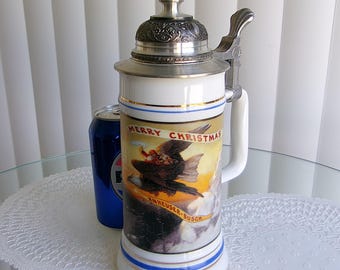 May include: A white ceramic beer stein with a silver metal lid and handle. The stein features a colorful illustration of an eagle carrying Santa Claus with the words "MERRY CHRISTMAS" and "Anheuser-Busch". A blue can is next to the stein.