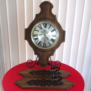 May include: A wooden wall clock with a silver face and black Roman numerals, displayed on a black metal stand. Below the clock are two wooden decorative door pulls with ornate black metal handles. The clock and pulls are on a red surface.