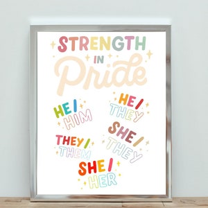 May include: A framed print featuring the words "STRENGTH IN Pride" in bright, playful lettering. Pronouns including "HE/HIM", "SHE/HER", and "THEY/THEM" are displayed in various colours. The print is displayed on a wooden surface.