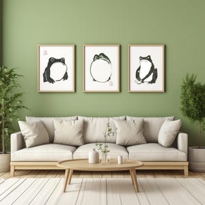 May include: Three framed black and white ink drawings of frogs on a light green wall above a white sofa with two cushions. The sofa is in front of a wooden coffee table with a vase of greenery and a white ceramic object.