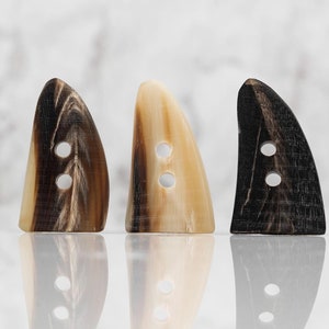 May include: Three horn buttons in different shades of brown. The buttons are shaped like a shark fin with two holes for sewing.