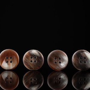 Puede incluir: Cuatro botones de cuerno marrón y blanco con cuatro agujeros cada uno. Los botones están dispuestos en una fila sobre un fondo negro.