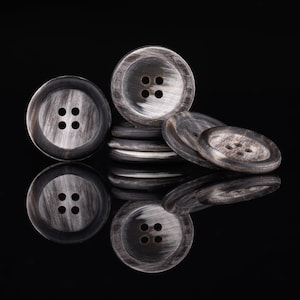 May include: A close-up of a pile of black and white horn buttons. The buttons are round and have four holes. The buttons are arranged in a pile, with the largest button on top.
