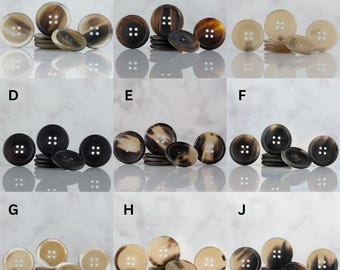 May include: A collection of various-sized buttons in shades of brown, tan, and cream. Each button has four holes and a slightly domed surface. The buttons are arranged in groups, with larger and smaller sizes.