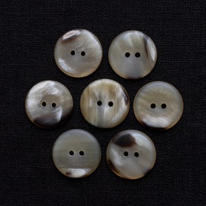May include: Seven round, light-colored buttons with two holes each, arranged in a circular pattern. The buttons have a marbled appearance with shades of brown and cream. They are set against a dark, textured background.