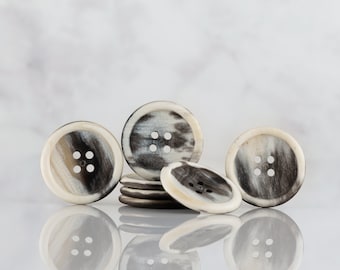 May include: A collection of round buttons with four holes each. The buttons have a cream-colored outer ring and a center with a mix of black, brown, and cream patterns. The buttons are arranged on a reflective surface.