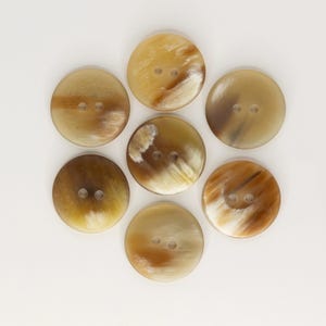 May include: A collection of seven round buttons with two holes each, arranged in a circular pattern. The buttons have a natural, mottled appearance with shades of brown, tan, and cream, suggesting a horn or similar material. The buttons are set against a white background.