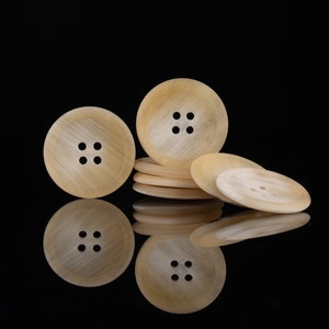 May include: A group of four beige buttons with four holes each. The buttons are arranged in a stack with one button on top of the others. The buttons are on a black surface.