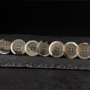 Può includere: Sette bottoni rotondi con quattro fori ciascuno, disposti in fila. I bottoni hanno un colore marrone chiaro e crema con un effetto marmorizzato. Sono posizionati su uno sfondo scuro e strutturato.