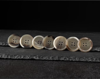 May include: Seven round buttons with four holes each, arranged in a row. The buttons have a light brown and cream color with a marbled effect. They are set against a dark, textured background.