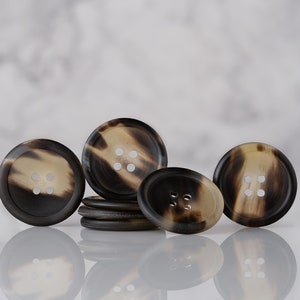 May include: Four brown and beige horn buttons with four holes each. The buttons are arranged in a semi-circle on a white surface.