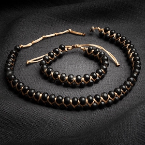 May include: A black beaded necklace and bracelet set. The round beads are strung together with a gold-coloured cord. The jewellery is displayed on a dark grey fabric.