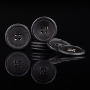 Puede incluir: Un grupo de botones negros con cuatro agujeros cada uno, dispuestos en una pila sobre una superficie negra.