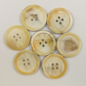 Peut inclure: Sept boutons ronds de couleur crème avec quatre trous chacun. Les boutons sont en corne et présentent un motif naturel marbré.