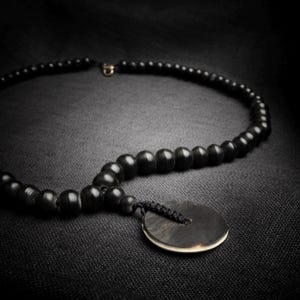 May include: A black beaded necklace with a circular pendant. The necklace features dark, round beads and a pendant with a light-colored edge. The necklace is displayed on a dark, textured surface.