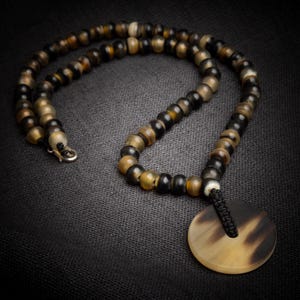 May include: A beaded necklace with a circular pendant. The necklace features a mix of brown, black, and tan beads. The pendant is a flat, circular piece with a dark brown and tan pattern. The necklace is displayed on a dark gray textured surface.