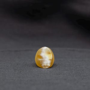 May include: A smooth, rounded ring featuring a translucent golden-yellow hue with a distinct, shimmering white chatoyant band running vertically through the center, creating a cat's eye effect within the polished, solid material of the band.