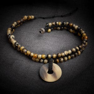 May include: A handmade necklace with a circular pendant and a beaded design. The necklace features small, round beads in shades of brown, tan, and black. The pendant is light brown with a dark brown pattern. The necklace is displayed on a dark grey surface.