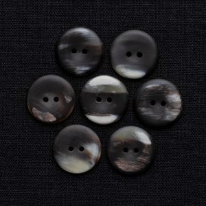 May include: Seven round buttons with two holes each, arranged in a circular pattern on a black fabric. The buttons have a marbled appearance with shades of black, brown, and cream, creating a natural, textured look. The buttons are likely for sewing or crafts.