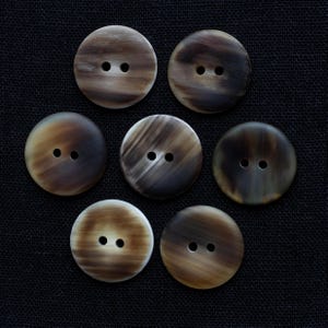 May include: Seven round buttons with two holes each, in shades of brown and tan. The buttons have a marbled appearance, with swirling patterns and variations in color. The buttons are arranged on a dark background.