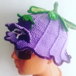 May include: Purple crocheted hat shaped like a flower with green leaves. The hat has a ruffled edge and a flower detail on the side.
