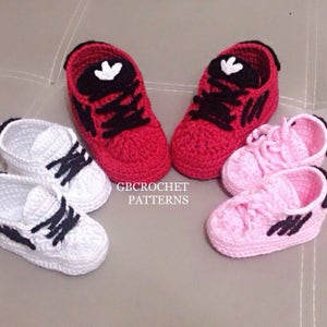May include: Three pairs of crocheted baby shoes in red, white and pink. The shoes have black laces and a white logo on the side. The shoes are arranged in a row on a light grey surface. GBcrochet Patterns is written below the shoes.