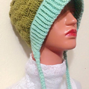 May include: A green and light blue crocheted hat with long ties. The hat has a unique design with a textured pattern.