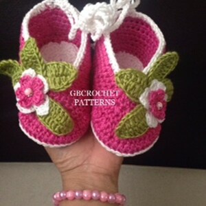 May include: A pair of pink crocheted baby booties with white and green flower accents. The booties have white laces and are tied together.