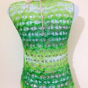May include: A sleeveless, open-weave crochet top in shades of green, transitioning from light to dark. The top features a chevron pattern and a halter neck tie. The neckline is adorned with a small, colourful beaded trim.