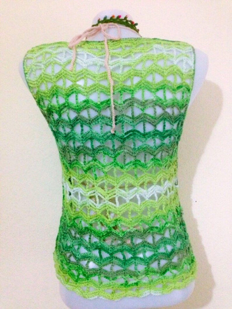 May include: A sleeveless, open-weave crochet top in shades of green, transitioning from light to dark. The top features a chevron pattern and a halter neck tie. The neckline is adorned with a small, colourful beaded trim.