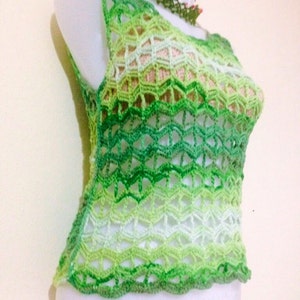 May include: A sleeveless, green crochet top with a scalloped hem. The top features a gradient of green shades, from light to dark, creating a textured, open-weave design. A matching green and floral necklace is also visible.