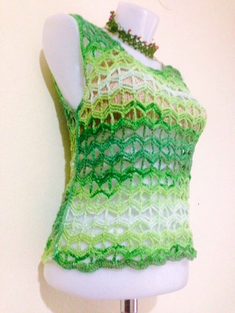 May include: A sleeveless, green crochet top with a scalloped hem. The top features a gradient of green shades, from light to dark, creating a textured, open-weave design. A matching green and floral necklace is also visible.