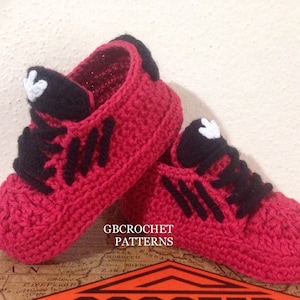 May include: A pair of red crocheted baby shoes with black laces and white accents. The shoes have a simple design and are perfect for a baby's first steps.